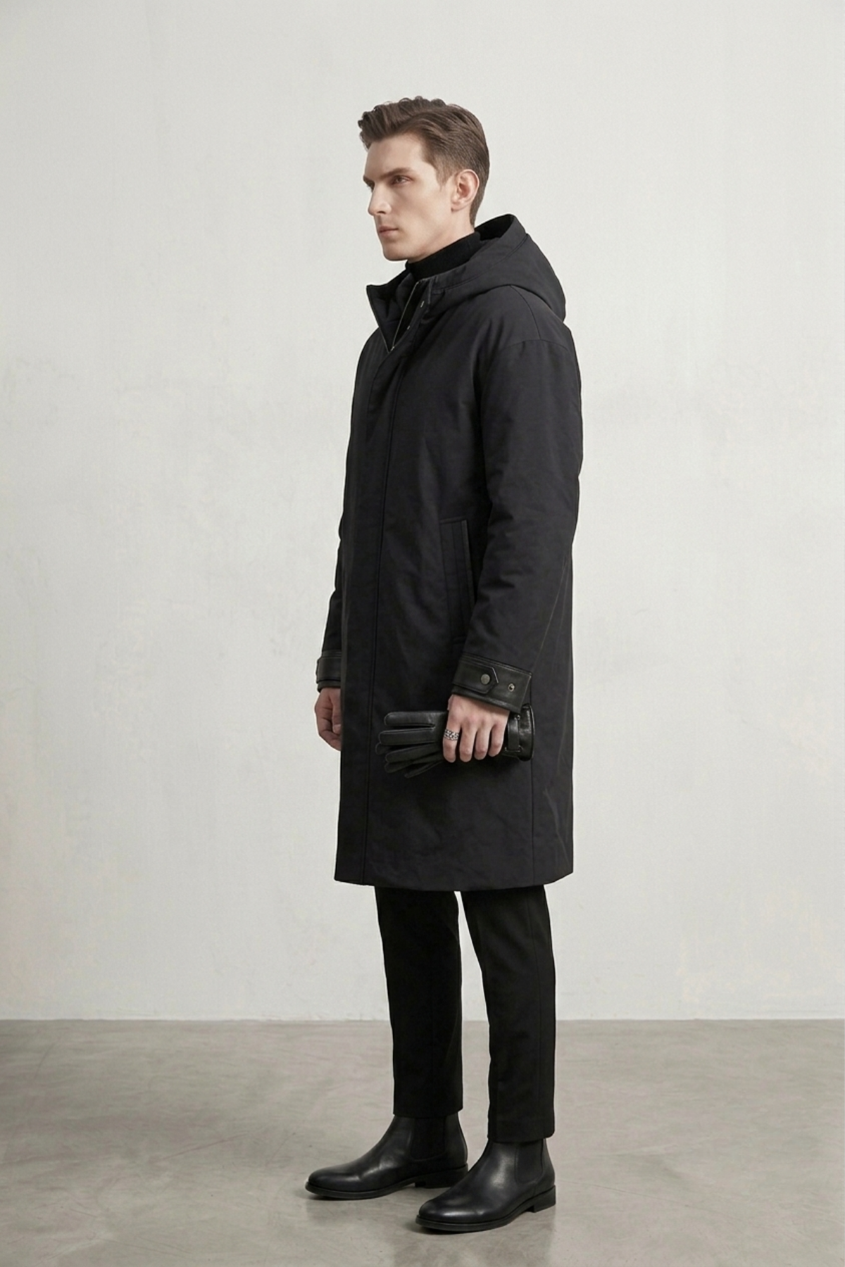 A man wearing a long black hooded coat, standing in profile against a light gray studio wall.