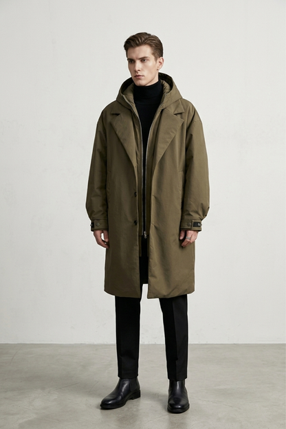 A man wearing a mid-length olive green hooded coat over a black turtleneck standing against a plain light gray studio background.